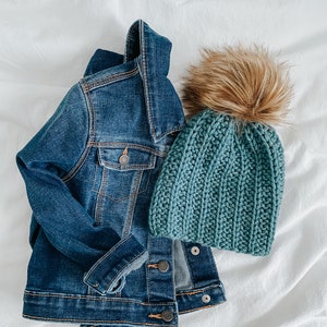May include: A blue denim jacket and a teal knitted beanie with a brown faux fur pom pom.