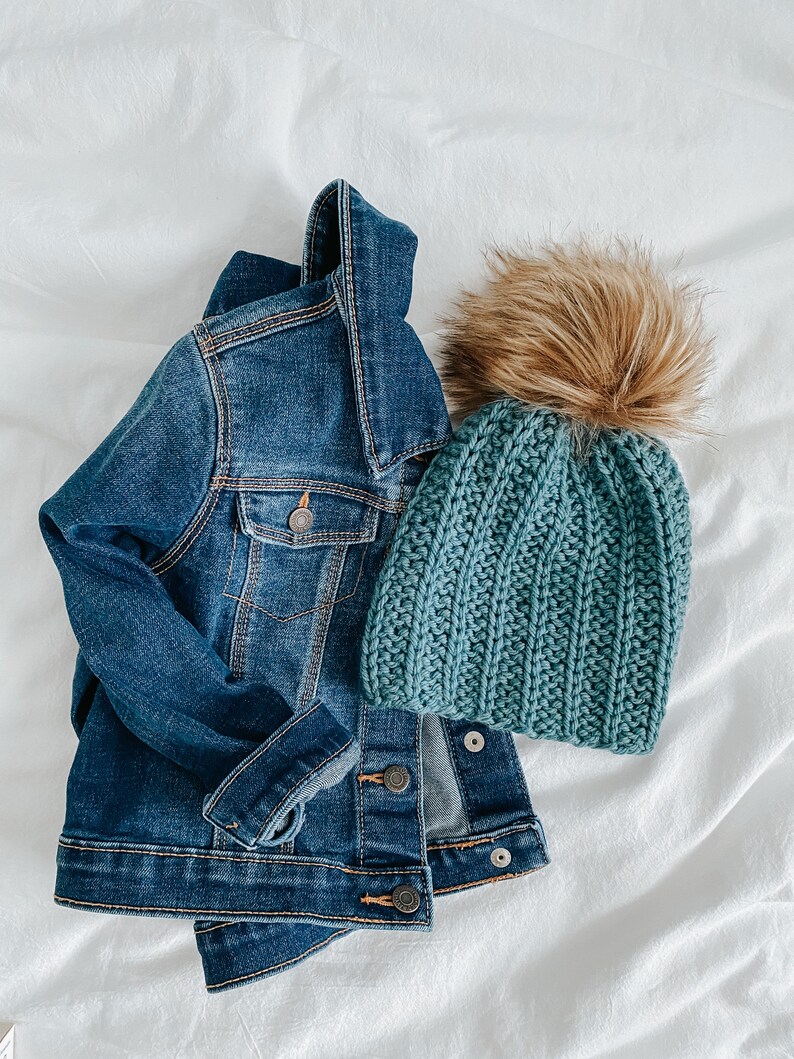 May include: A blue denim jacket and a teal knitted beanie with a brown faux fur pom pom.