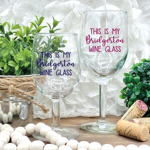 May include: Two clear wine glasses with the text "THIS IS MY Bridgerton WINE GLASS" in blue and pink lettering. The glasses are on a wooden surface with decorative elements, including corks and greenery. A charming gift for wine lovers.