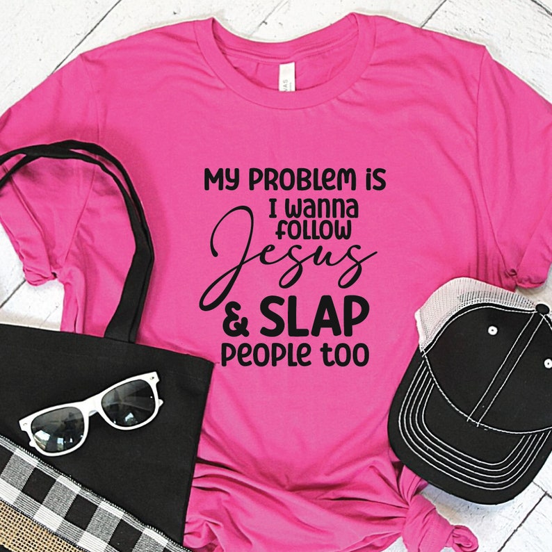 K&ouml;nnte beinhalten: Ein leuchtend pinkes Kurzarm-T-Shirt mit dem schwarzen Text: MY PROBLEM IS I WANNA FOLLOW Jesus & SLAP PEOPLE too. Das T-Shirt wird zusammen mit einer schwarzen Tragetasche, einer wei&szlig; gerahmten Sonnenbrille und einer schwarz-wei&szlig;en Netz-Baseballkappe gezeigt.