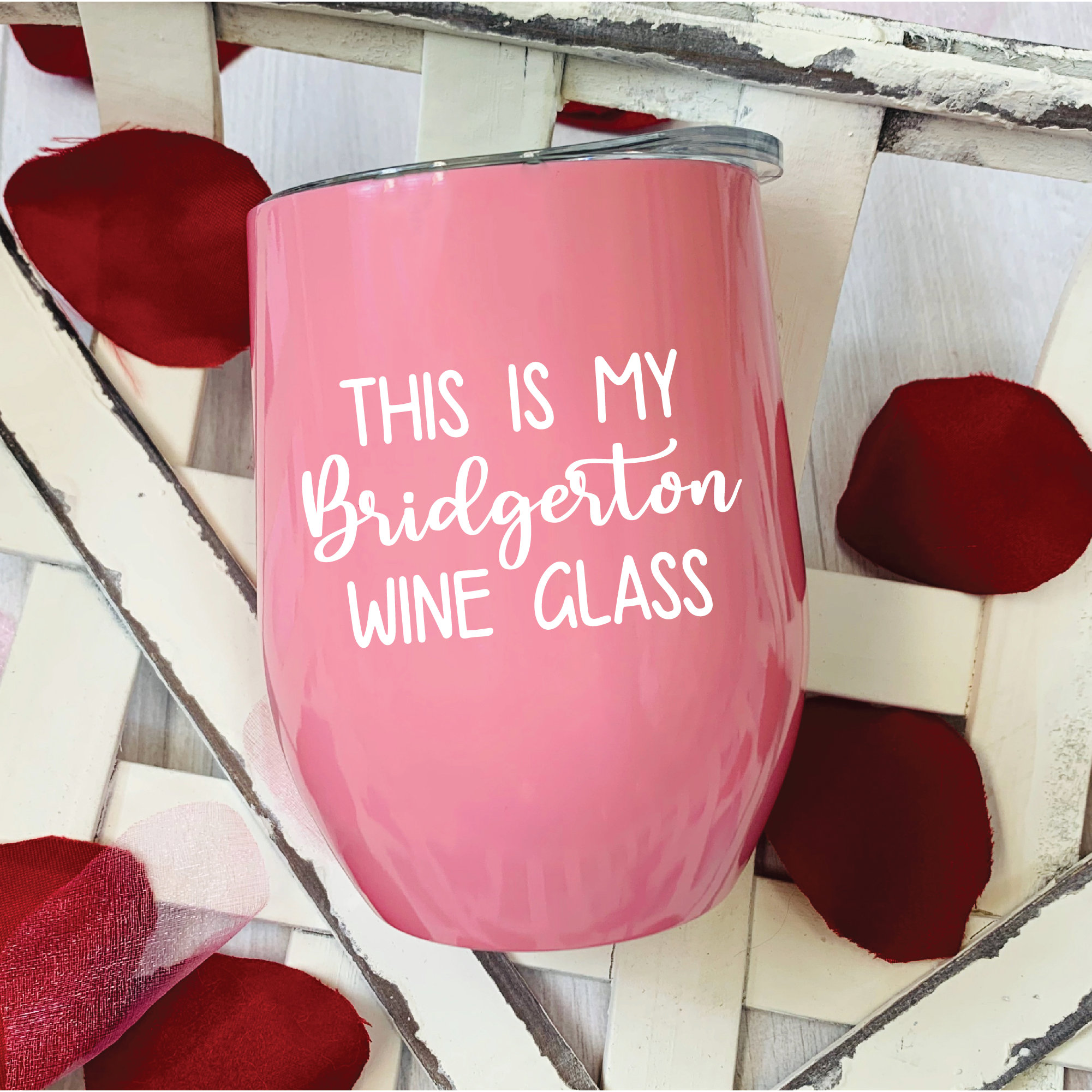May include: A pink stemless wine glass with a silver rim and a white lid. The text "THIS IS MY Bridgerton WINE GLASS" is printed on the front. Red rose petals are scattered around the glass.