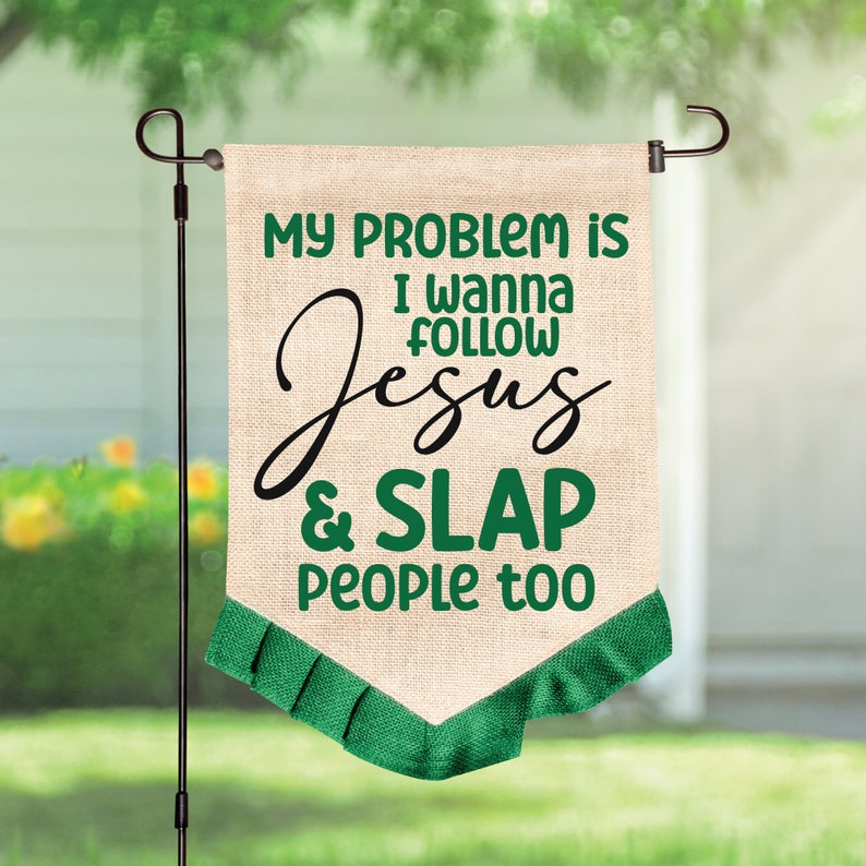 K&ouml;nnte beinhalten: Eine beige Gartenflagge in Jute-Optik mit gr&uuml;nem und schwarzem Text, der lautet: MY PROBLEM IS I WANNA FOLLOW JESUS & SLAP PEOPLE too. Die Flagge hat einen spitz zulaufenden unteren Rand mit einer plissierten gr&uuml;nen Stoffborte.