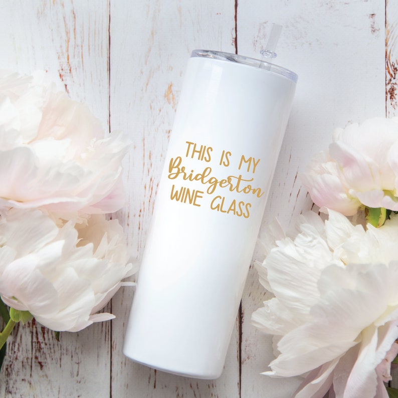 May include: A white tumbler with a clear straw and lid, featuring the text "THIS IS MY Bridgerton WINE GLASS" in gold lettering. The tumbler is surrounded by white and pink flowers, set against a light wood background.