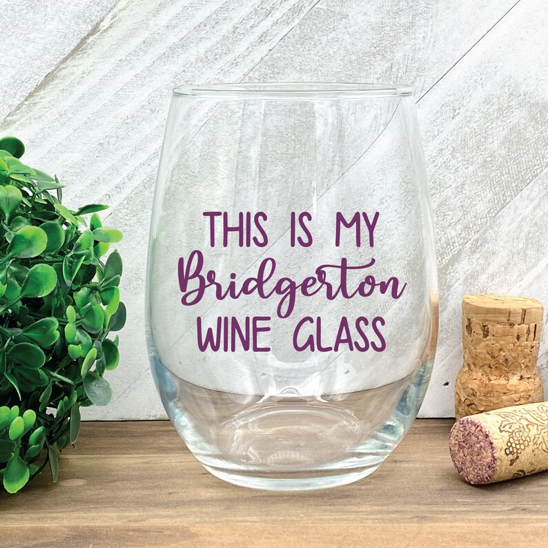 May include: A clear stemless wine glass with the text "THIS IS MY Bridgerton WINE GLASS" in purple script. A cork and a wine cork are next to the glass. The glass sits on a wooden surface.