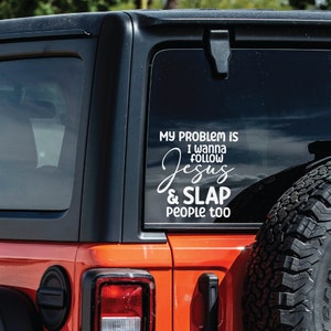 K&ouml;nnte beinhalten: Ein wei&szlig;er Vinyl-Aufkleber auf der Heckscheibe eines orangefarbenen Fahrzeugs. Der Aufkleber zeigt den Text: MY PROBLEM IS I WANNA FOLLOW JESUS & SLAP PEOPLE TOO. Die Schrift kombiniert eine serifenlose Druckschrift mit einer Schreibschrift.