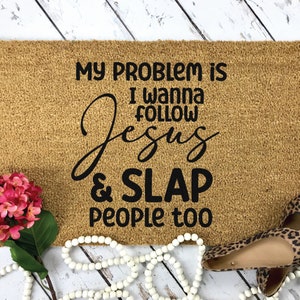 K&ouml;nnte beinhalten: Eine rechteckige, beigefarbene Kokosfasermatte mit schwarzem Text: MY PROBLEM IS I WANNA FOLLOW JESUS & SLAP PEOPLE too. Die Matte ist mit einem Strau&szlig; rosa Hortensien, einer wei&szlig;en Holzperlenkette und Schuhen mit Leopardenmuster verziert.