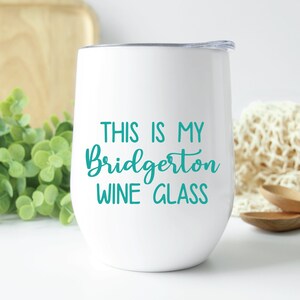 May include: White stemless wine glass with a teal inscription that reads "THIS IS MY Bridgerton WINE GLASS". The glass has a silver rim and a clear lid. The background includes greenery, a wooden cutting board, and a cream-colored cloth.