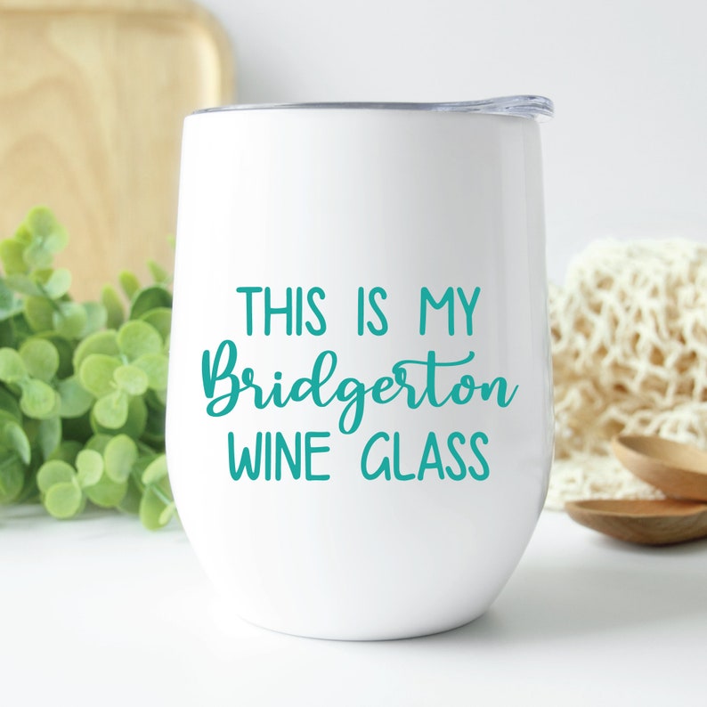May include: White stemless wine glass with a teal inscription that reads "THIS IS MY Bridgerton WINE GLASS". The glass has a silver rim and a clear lid. The background includes greenery, a wooden cutting board, and a cream-colored cloth.