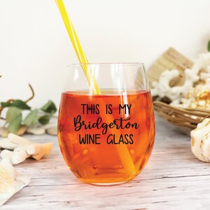 May include: A stemless wine glass filled with an orange liquid and a yellow and white striped straw. The glass has the text "THIS IS MY Bridgerton WINE GLASS" in black script. The glass is on a white wooden surface.