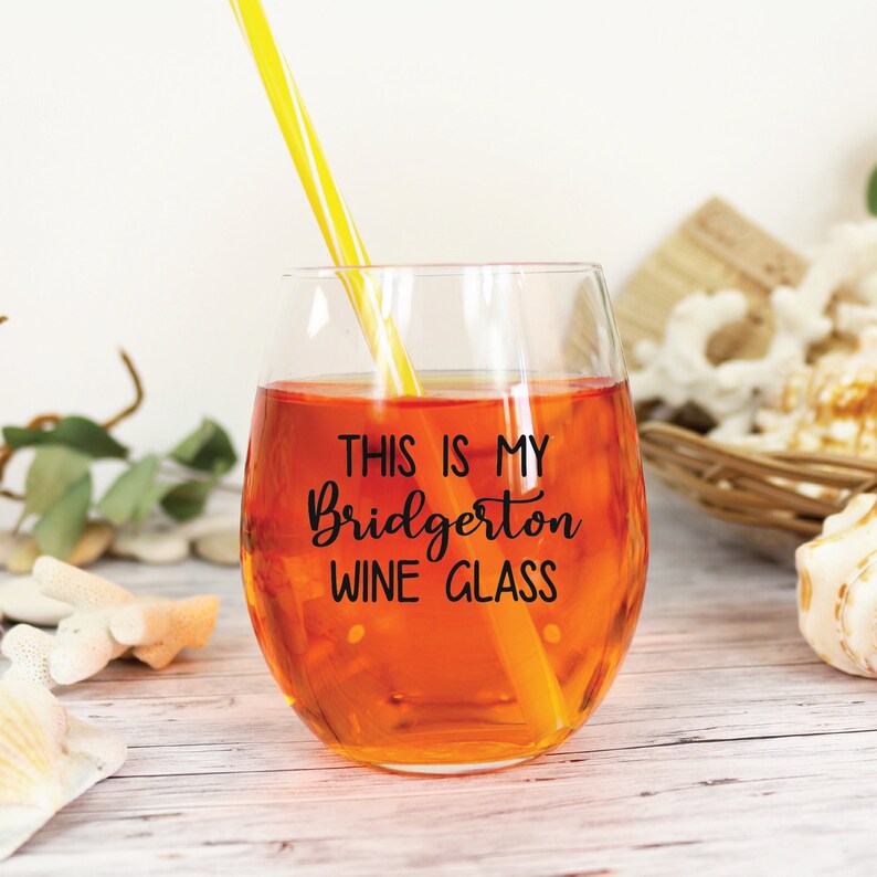 May include: A stemless wine glass filled with an orange liquid and a yellow and white striped straw. The glass has the text "THIS IS MY Bridgerton WINE GLASS" in black script. The glass is on a white wooden surface.