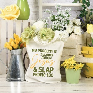 K&ouml;nnte beinhalten: Eine cremefarbene Stofftasche mit dem dunkelgr&uuml;nen und gelben Text "MY PROBLEM IS I WANNA FOLLOW JESUS & SLAP PEOPLE TOO". Die Tasche hat zwei Tragegriffe und ist mit einer Vielzahl von wei&szlig;en, gr&uuml;nen und gelben Kunstblumen gef&uuml;llt.
