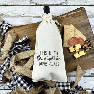 May include: A beige wine bottle bag with the text "THIS IS MY BRIDGERTON WINE GLASS" in black script. The bag is tied at the top and sits on a wooden cutting board with crackers, cheese, salami, and walnuts. Burlap and black and white checkered ribbon surround the board.