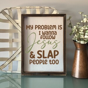 K&ouml;nnte beinhalten: Ein rechteckiges Holzschild mit wei&szlig;em Hintergrund und dunkelbraunem Rahmen. Das Schild tr&auml;gt braunen und salbeigr&uuml;nen Text mit der Aufschrift: MY PROBLEM IS I WANNA FOLLOW JESUS & SLAP PEOPLE TOO. Die Schriftart ist eine Mischung aus Block- und Schreibschrift.