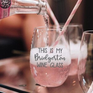 May include: A stemless wine glass filled with pink liquid and bubbles, with a pink and white striped straw. The glass has the text "THIS IS MY Bridgerton WINE GLASS" in a script font. A bottle of pink lemonade is pouring into the glass.