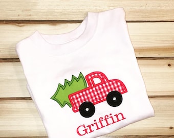 May include: White t-shirt with a red and white gingham truck carrying a green Christmas tree. The name "Griffin" is embroidered in pink below the truck.