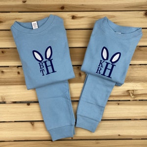 May include: Two sets of light blue pajamas with embroidered bunny ears and initials. Each set includes a long-sleeved top and matching pants. The bunny ears are white, and the initials are dark blue. The pajamas are laid out on a wooden surface.