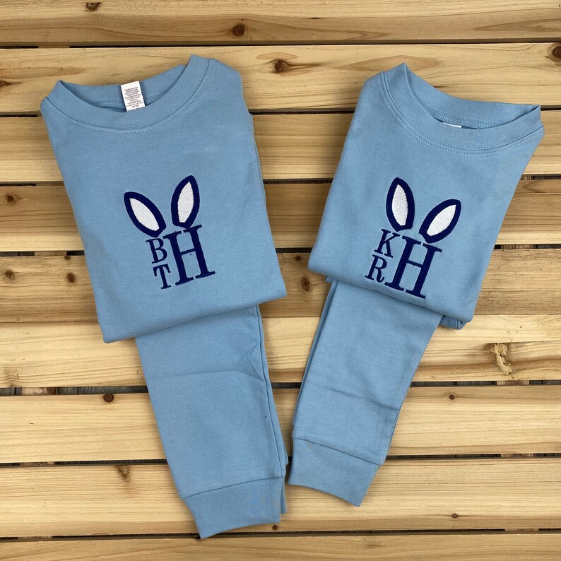 May include: Two sets of light blue pajamas with embroidered bunny ears and initials. Each set includes a long-sleeved top and matching pants. The bunny ears are white, and the initials are dark blue. The pajamas are laid out on a wooden surface.