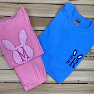 May include: Two children's shirts on a wooden surface. One is pink with a bunny face and the letter "M". The other is blue with bunny ears and the letters "RRR". Both shirts are long-sleeved.