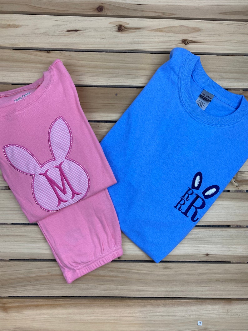 May include: Two children's shirts on a wooden surface. One is pink with a bunny face and the letter "M". The other is blue with bunny ears and the letters "RRR". Both shirts are long-sleeved.