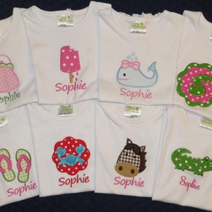 May include: Eight white t-shirts with different embroidered designs. Each shirt features a different design, including a purse, popsicle, whale, letter S, flip flops, crab, horse, and alligator. Each shirt has the name "Sophie" embroidered on it.