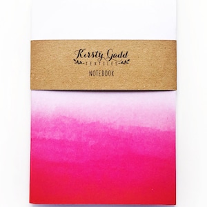 May include: A pink and red ombre notebook with a brown paper label that reads "Kirsty Gadd Textiles Notebook".