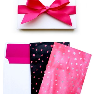 May include: A white gift box with a pink satin ribbon tied in a bow. The box is open and contains three envelopes with pink and black polka dot patterns.