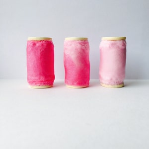 May include: Three spools of pink ribbon, each with a different shade of pink. The spools are arranged in a row from left to right, with the darkest pink spool on the left and the lightest pink spool on the right.