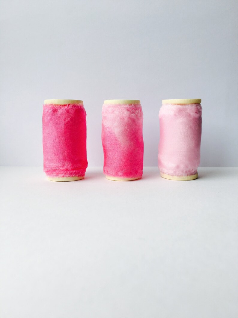 May include: Three spools of pink ribbon, each with a different shade of pink. The spools are arranged in a row from left to right, with the darkest pink spool on the left and the lightest pink spool on the right.