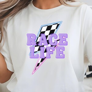 May include: White t-shirt featuring a purple, blue, and pink lightning bolt graphic with the words "RACE LIFE" in purple block letters. The lightning bolt includes a checkered flag design. The shirt is a casual, crew neck style.