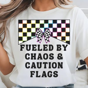 May include: White t-shirt with a graphic design featuring a checkered flag and the text "Fueled by Chaos & Caution Flags". The design includes a black and white checkered bow. The shirt is worn with blue jeans.