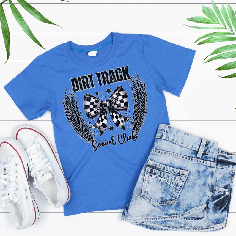 Pu&ograve; includere: Una maglietta blu con la scritta "DIRT TRACK Social Club" e un fiocco a scacchi. &Egrave; abbinata a pantaloncini di jeans e scarpe da ginnastica bianche con bordi rossi, il tutto su una superficie in legno bianco.