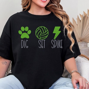 Pode incluir: Camiseta preta com desenhos de strass verdes de uma pegada, uma bola de voleibol e um raio. As palavras "DIG SET SPIKE" estão abaixo dos gráficos em letras de strass. A camisa é de estilo casual.