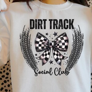 Pu&ograve; includere: Felpa bianca con un design grafico. Il design presenta il testo "DIRT TRACK Social Club", un fiocco a scacchi, stelle e dettagli di impronte di pneumatici, creando un tema di corse.
