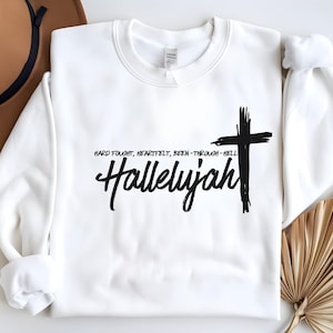 Może przedstawiać: Biała bluza z czarnym krzyżem i tekstem "Hard fought, heartfelt, been through hell Hallelujah".