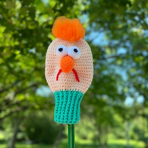 May include: A handmade golf club head cover in the shape of a cartoon character's face. The cover is primarily beige with orange hair, an orange nose, and red eyebrows. The bottom is green, and the eyes are white with black pupils.