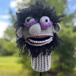 May include: A crocheted golf club head cover featuring a character with a gray face, purple eyelids, a black beard, and a wide, toothy grin.