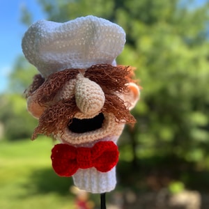 May include: Crocheted puppet head with a white chef's hat, brown mustache and eyebrows, a red bow tie, and an open mouth.