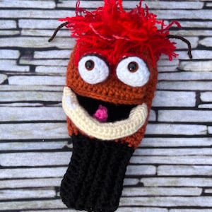 May include: Crocheted golf club head cover featuring a brown and orange monster with red hair, white eyes, and a big smile. The cover is black at the bottom.