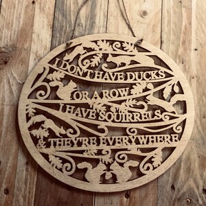 Può includere: Un cartello rotondo in legno con la scritta "I don't have ducks or a row, I have squirrels & they're everywhere." Il cartello presenta disegni di scoiattoli e foglie di quercia ritagliati. Un cordino di iuta è attaccato per appenderlo.