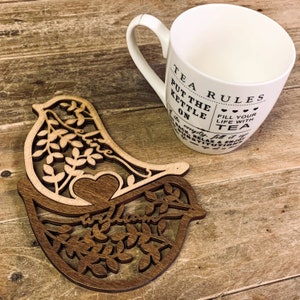 Puede incluir: Un posavasos de madera con forma de pájaro con un corazón recortado y diseños intrincados de hojas. El posavasos es marrón y tiene la palabra "creer" tallada en él. Una taza blanca con texto negro que dice "Tea Rules Put the Kettle On... Fill your life with tea" está sentada sobre una superficie de madera.