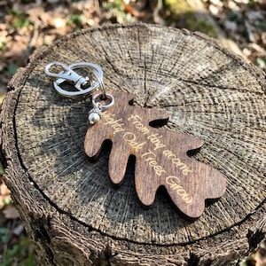 Könnte beinhalten: Ein hölzerner Schlüsselanhänger in Blattform mit dem eingravierten Text "From tiny acorns mighty Oak Trees Grow". Der Schlüsselanhänger hat einen silberfarbenen Verschluss und einen kleinen Eichel-Anhänger. Das Holz hat eine dunkelbraune Farbe.