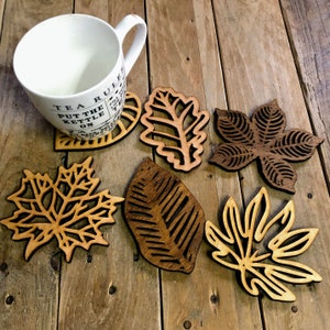 May include: A white ceramic mug with the text "TEA RULES PUT THE KETTLE ON" rests on a wooden leaf-shaped coaster. Several other wooden leaf coasters in various designs and shades of brown are scattered on a rustic wooden surface.