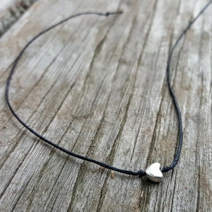 Here is a close-up of the heart wish bracelet. The heart metal bead is silver and flat and has a black waxed cotton cord going through the middle, out the top, and bottom. It is laying on a wood background.