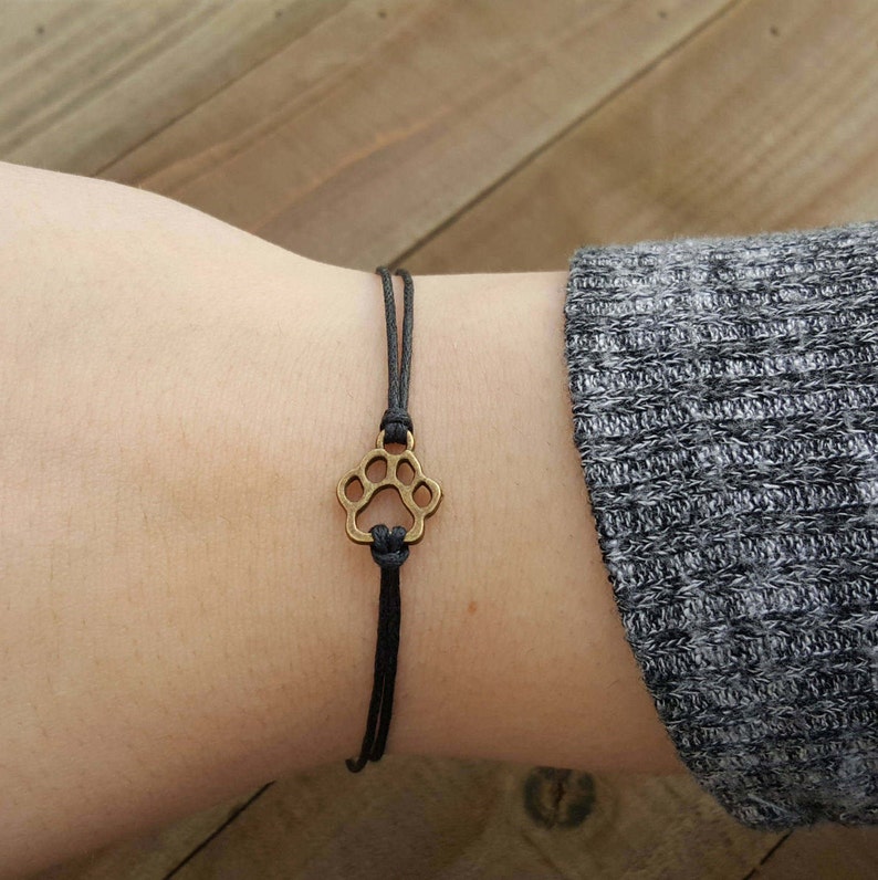 Here is a cute paw print wish bracelet. The paw print is bronzed and has black waxed cotton cord attached on the top and bottom. It is tied on to a wrist and the women has on a gray sweater.