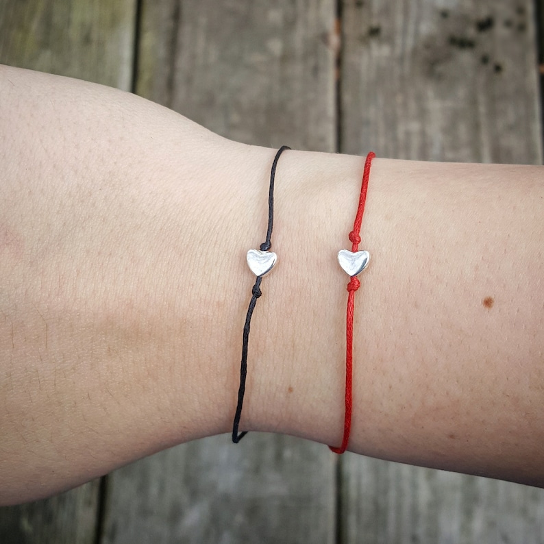 Here are two examples of the heart wish bracelets. The heart metal bead is silver and flat and has a red waxed cotton cord going through the middle, out the top, and bottom as well as one that has a black cord. They are tied on to a wrist.