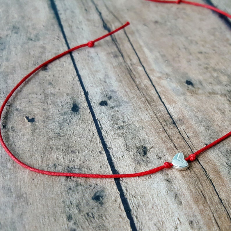 Here is a close up of the heart wish bracelet. The heart metal bead is silver and flat and has a red waxed cotton cord going through the middle, out the top, and bottom. It is laying on a wood background.