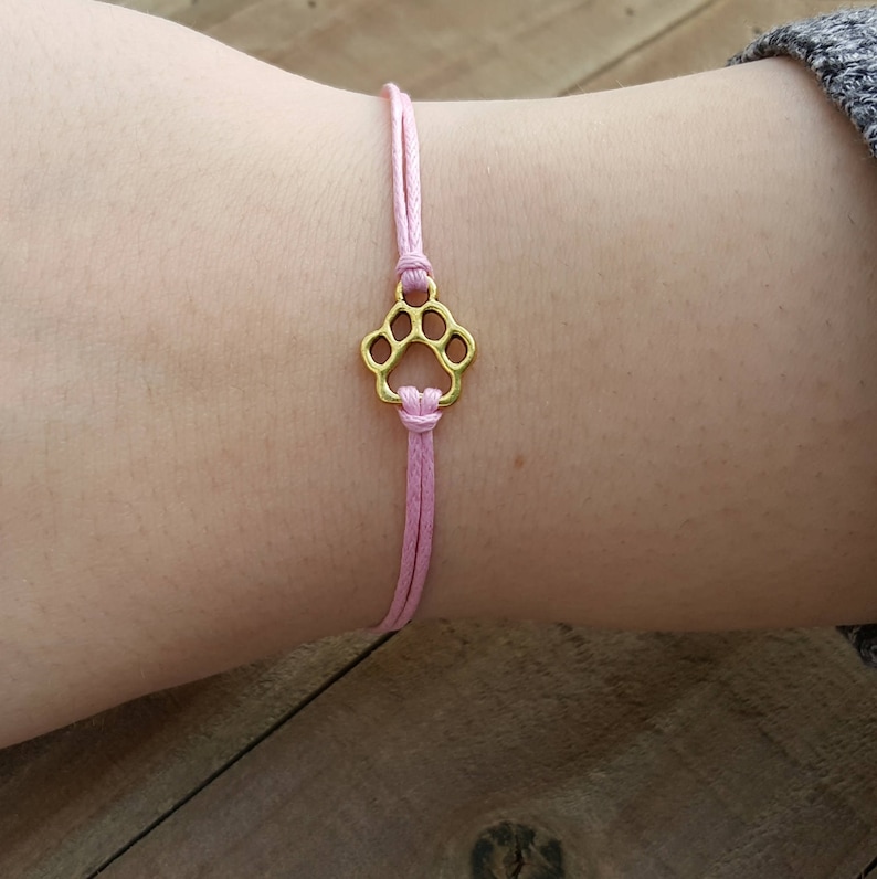 Here is a cute paw print wish bracelet. The paw print is golden and has a light pink waxed cotton cord attached on the top and bottom. It is tied on to a wrist and the women has on a gray sweater.