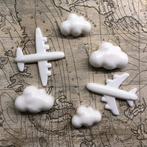 May include: White ceramic airplane and cloud shapes on a vintage world map background. The airplane shapes are detailed with wings, a tail, and a propeller. The cloud shapes are simple and rounded.