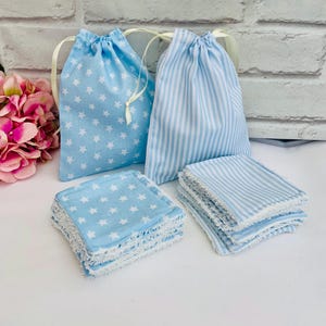 May include: Two light blue drawstring bags, one with white stars and the other with vertical stripes. Several matching square cloths are stacked nearby. The cloths and bags are made of fabric with a soft, textured appearance. The background includes a pink flower arrangement.