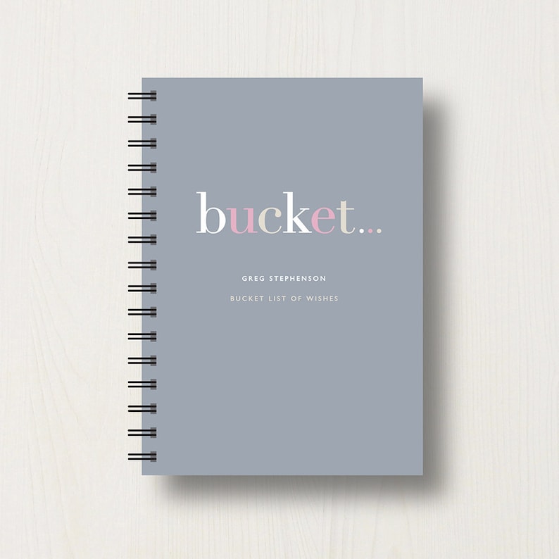 May include: A grey spiral bound notebook with the text "bucket..." in white and pink lettering. The text "GREG STEPHENSON" and "BUCKET LIST OF WISHES" is printed in white below the title.